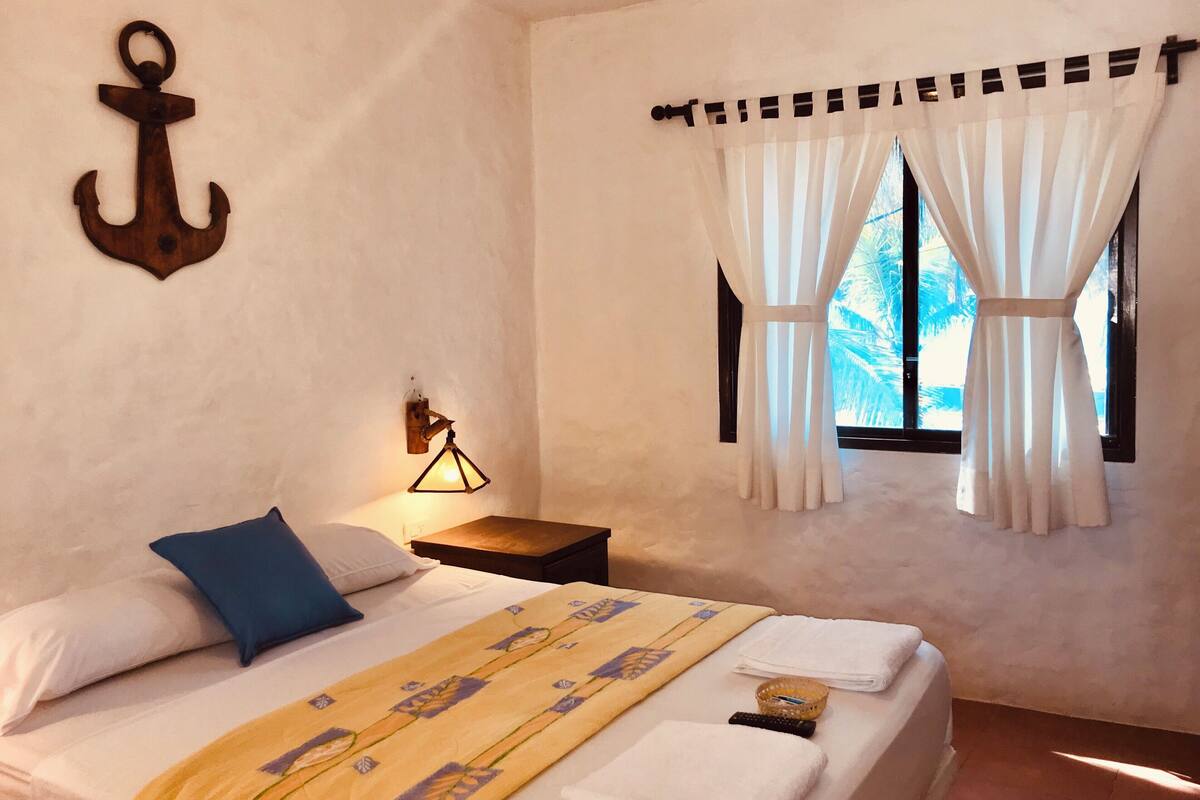 A bedroom with a bed, pillows, a bedside lamp, a window with curtains, and an anchor decoration on the wall.