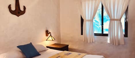 A bedroom with a bed, pillows, a bedside lamp, a window with curtains, and an anchor decoration on the wall.