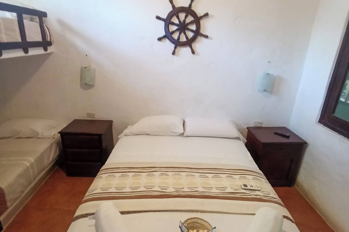 A single bed with a decorative headboard, a wall-mounted ship's wheel, and a window with a view.