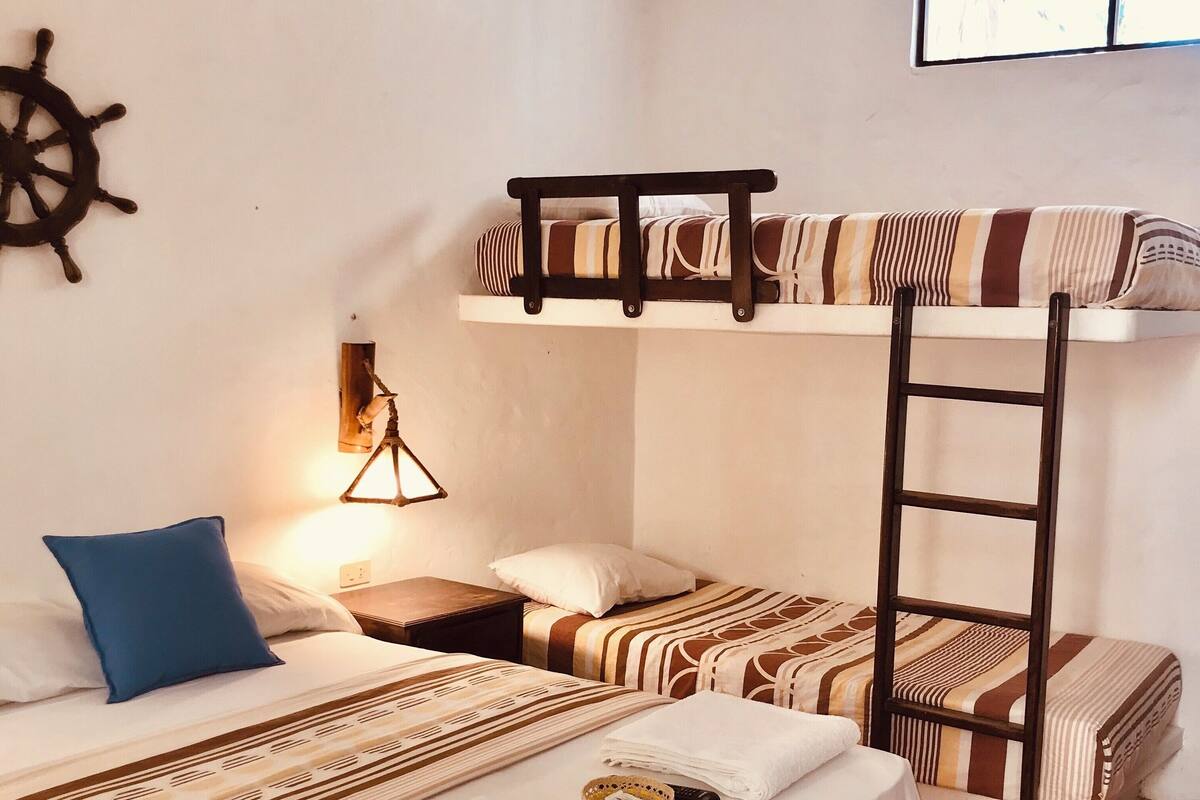 A dormitory room with bunk beds, a ship's wheel decoration, a wall-mounted lamp, and a window with blinds.