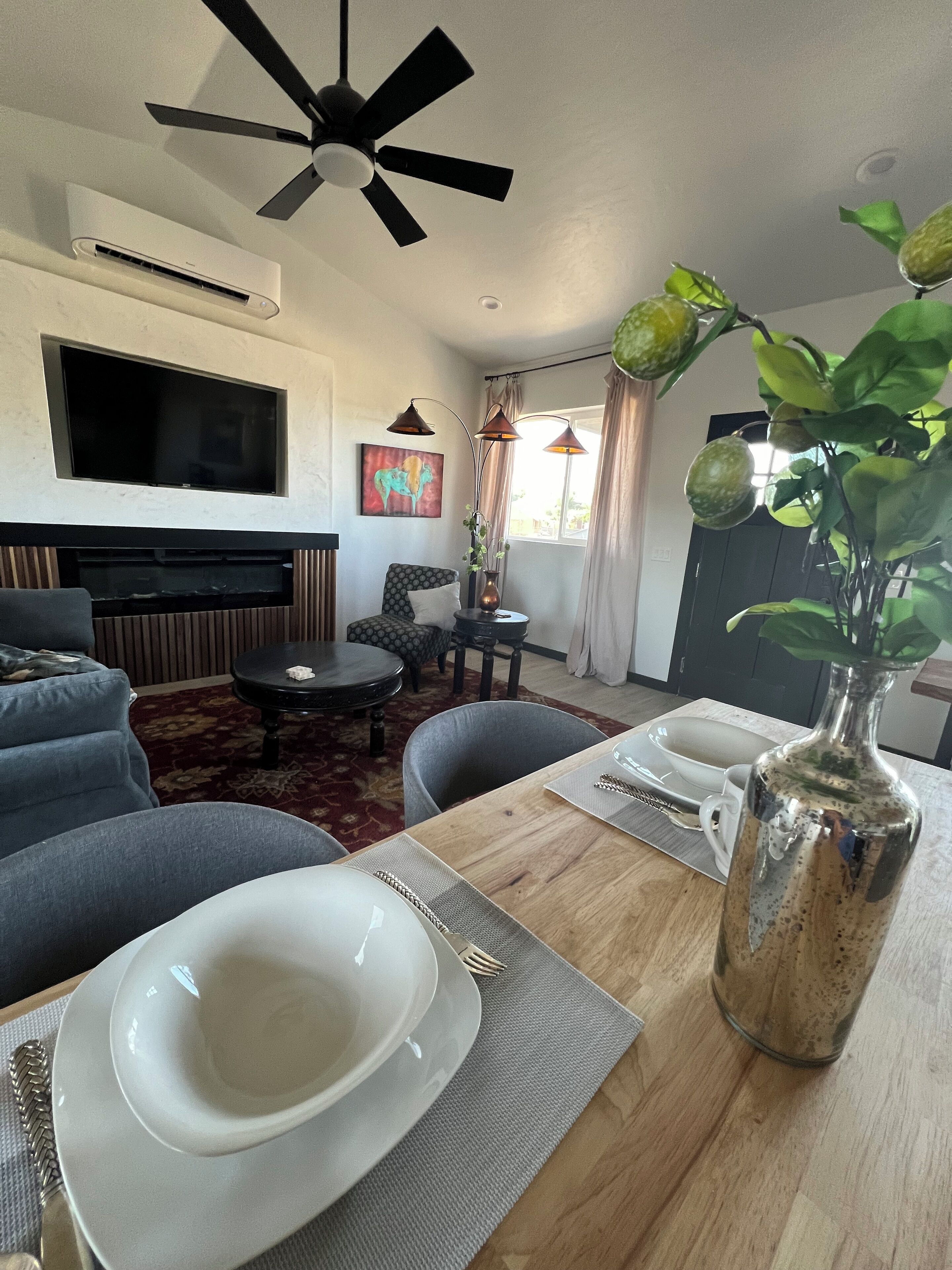 Un salon équipé d’un téléviseur à écran plat, d’un ventilateur de plafond, d’une table à manger dressée avec des assiettes, d’un vase avec une plante et d’un canapé.