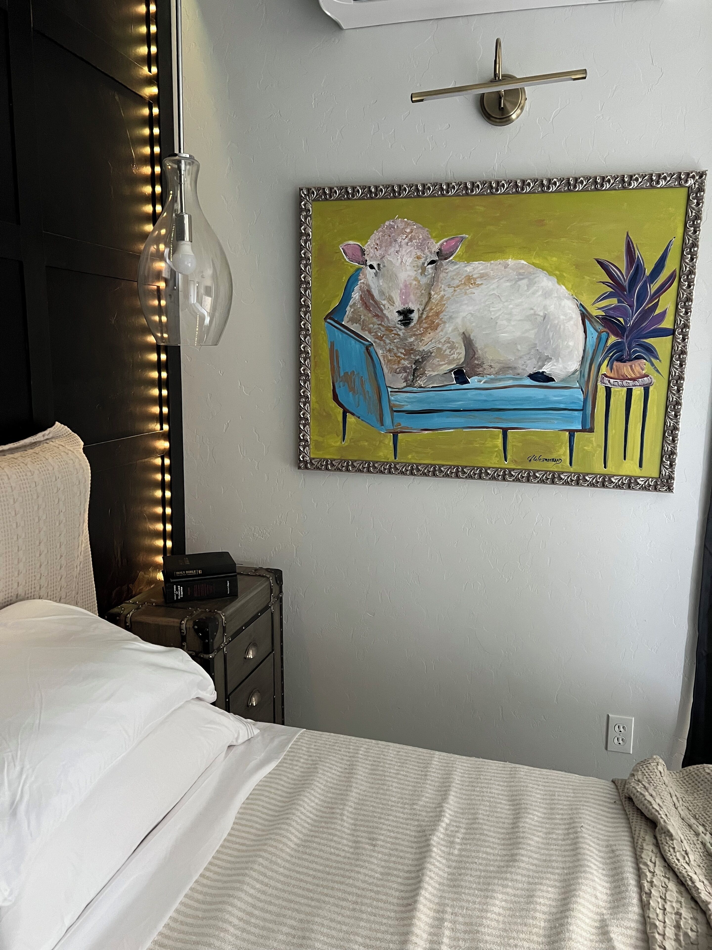 A bedroom with a bed, a bedside table with a lamp, a framed painting of a sheep on a couch, and a wall-mounted light fixture.