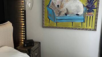 A bedroom with a bed, a bedside table with a lamp, a framed painting of a sheep on a couch, and a wall-mounted light fixture.