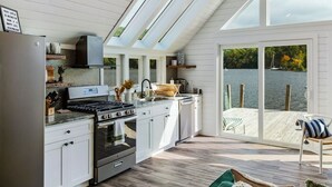 A modern kitchen with a large island, built-in appliances, and a sliding glass door leading to a deck with a view of a lake. - Dockside Dream | 4 mins to Mt Sunapee | Dock | VIEWS | Grill (Newbury)