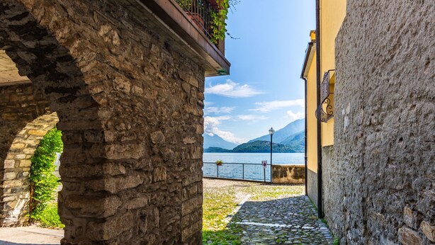 A narrow stone pathway leading to a scenic lake view, flanked by stone buildings with balconies.