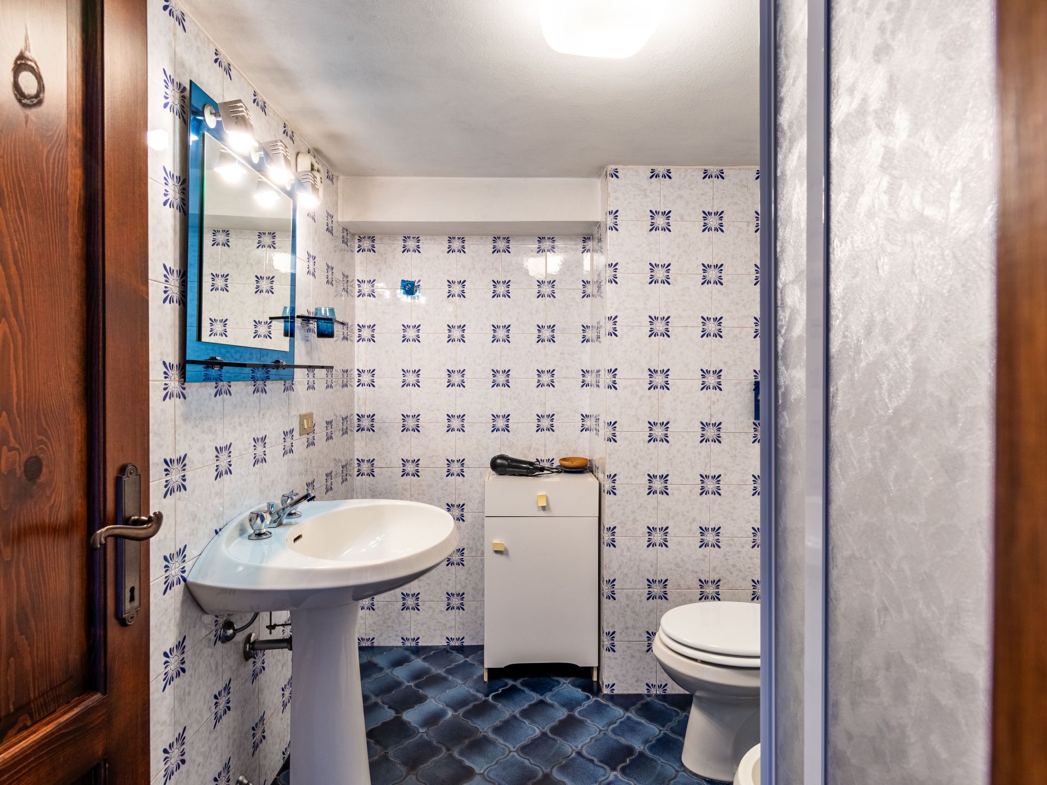 Une salle de bain avec un lavabo blanc, un miroir et des toilettes. Les murs sont recouverts de carreaux bleus à motifs floraux.