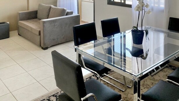 A modern living room with a glass-top dining table, black chairs, a grey sofa, and a potted orchid. - Pérola do Aquarius IV (São José dos Campos)