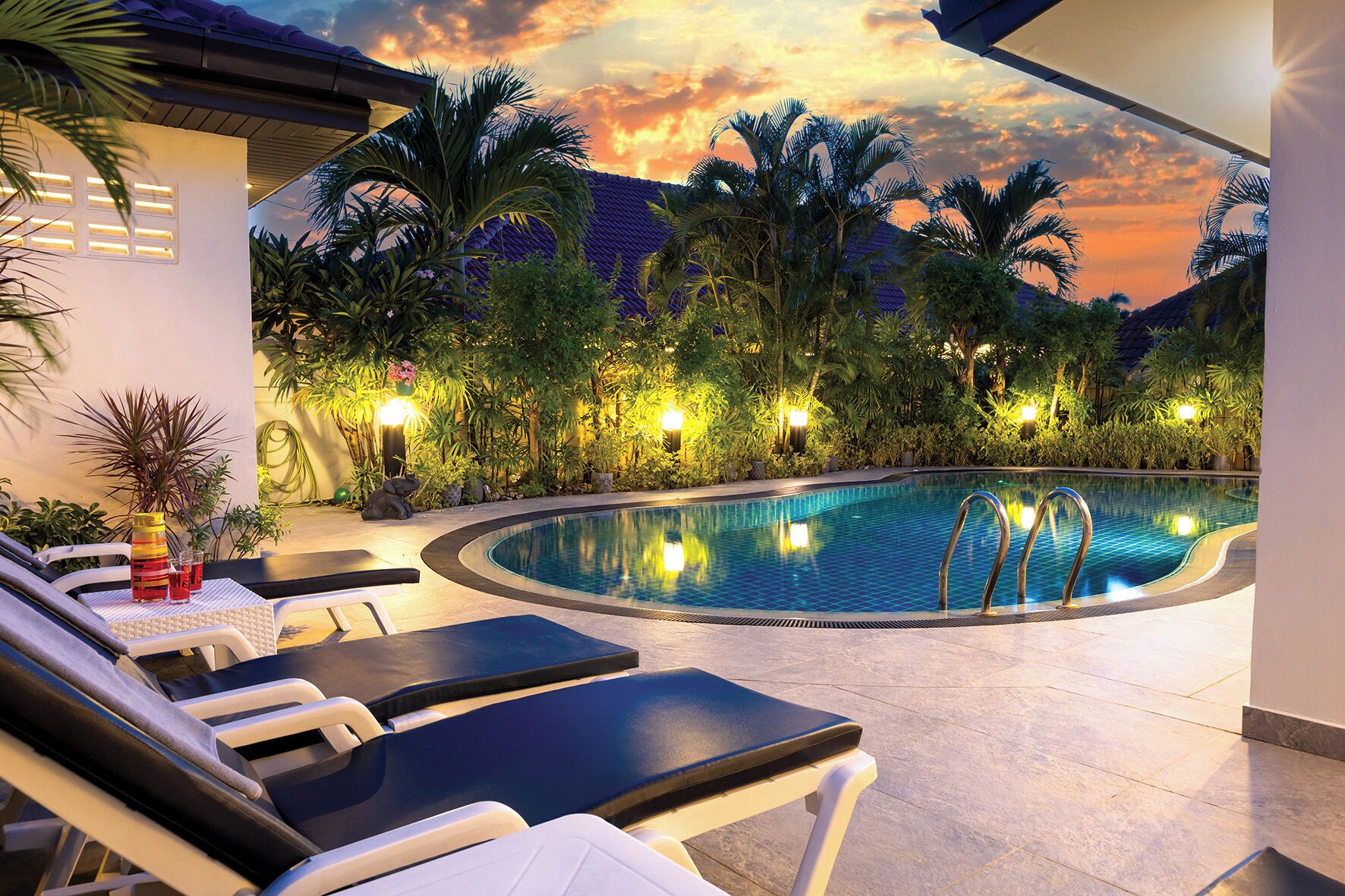 Uma piscina com espreguiçadeiras, rodeada por plantas tropicais e luzes noturnas.
