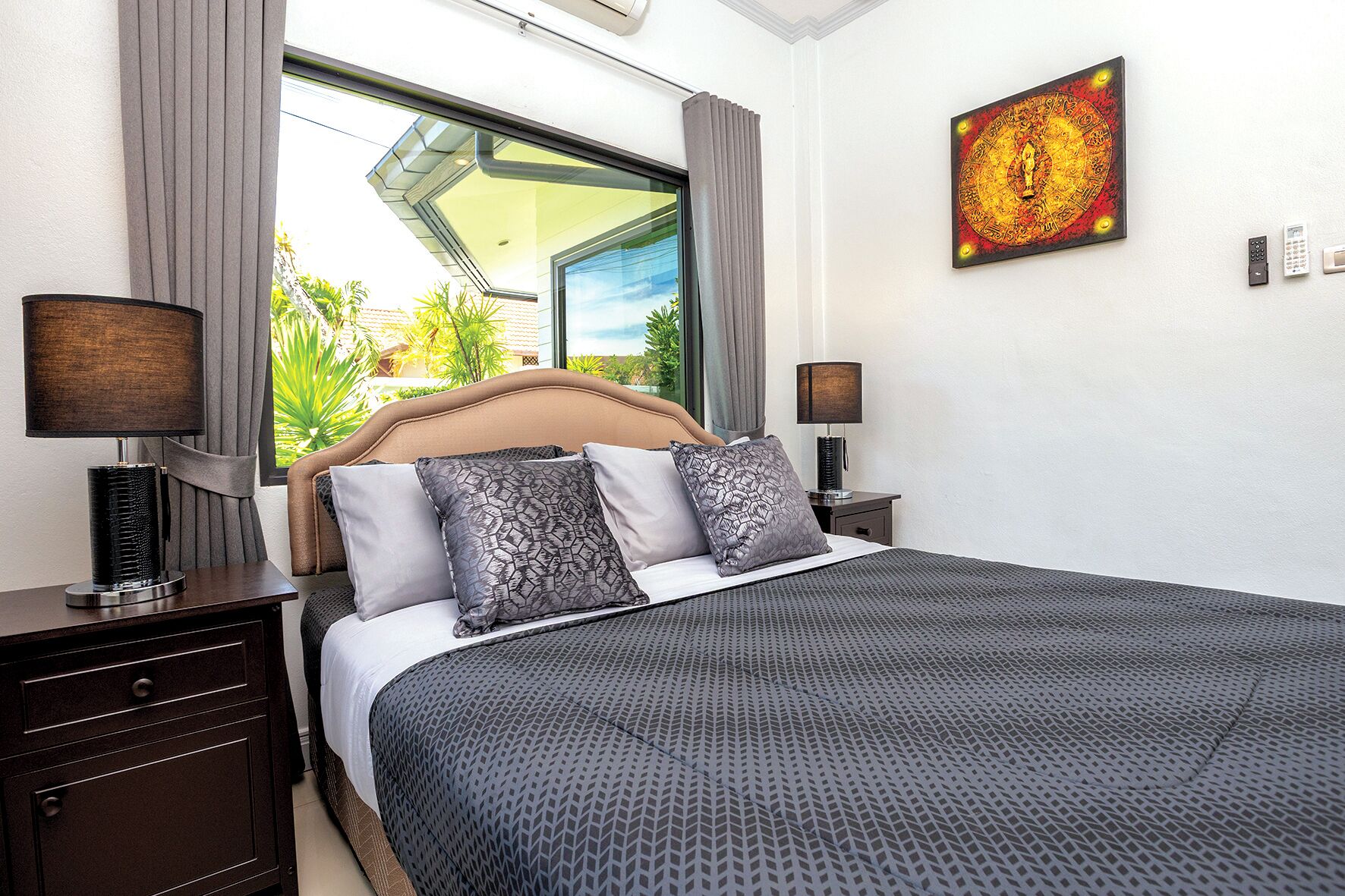 A hotel room with a large bed, two bedside tables, a window with curtains, a wall-mounted artwork, and a view of greenery outside.