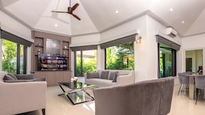 Ein geräumiges Wohnzimmer mit einer Couch, Sesseln, einem Couchtisch und einem Fernseher. Es gibt eine Küchenzeile mit einem Kühlschrank sowie einen Esstisch mit Stühlen. Große Fenster bieten Blick ins Grüne.