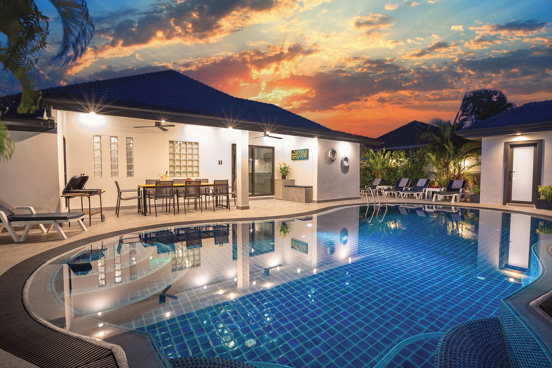 A swimming pool with lounge chairs and a dining area, surrounded by buildings with a sunset view.