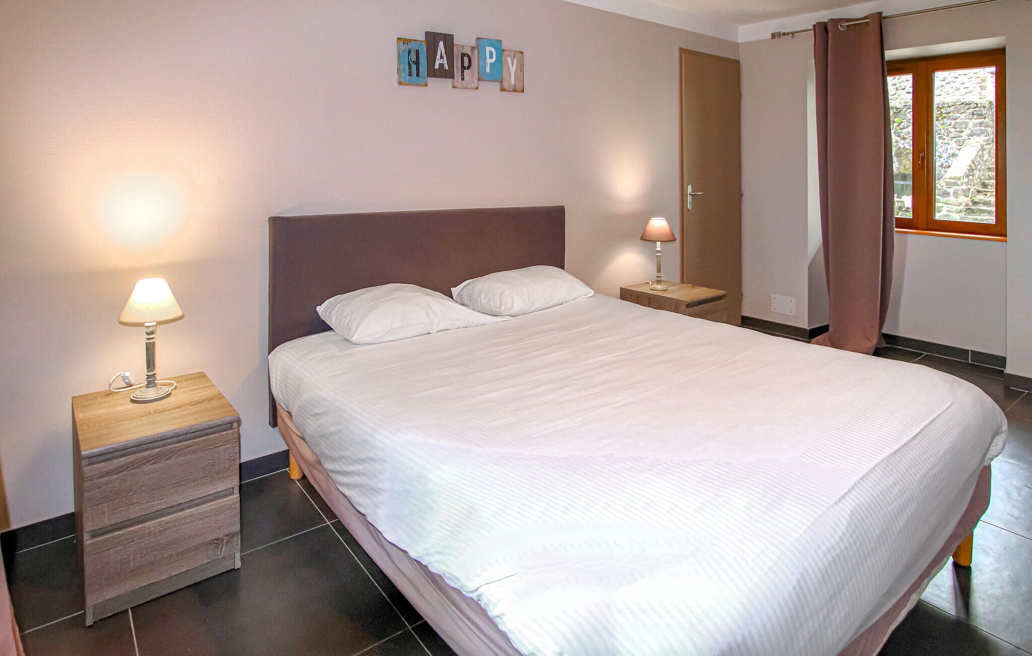 Een net opgemaakt bed in een hotelkamer, met een hoofdeinde, twee nachtkastjes met lampen, een klein houten dressoir en een 'HAPPY' decoratie aan de muur.