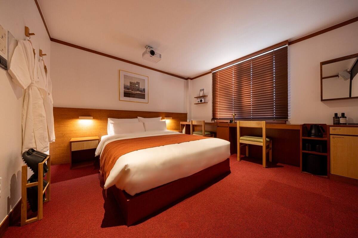 A hotel room with a large bed, wooden headboard, a desk with a chair, a small shelf with toiletries, and a window with blinds.