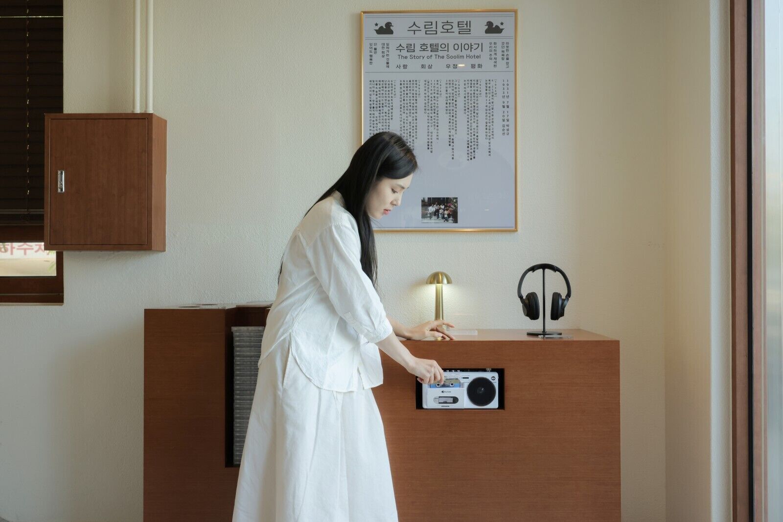 A person standing at a wooden reception desk with a radio, headphones, and a framed picture on the wall.