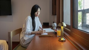Una persona escribiendo en un escritorio de madera con una lámpara, un vaso de jugo de naranja y un ventilador en una habitación con ventana y televisor en la pared.
