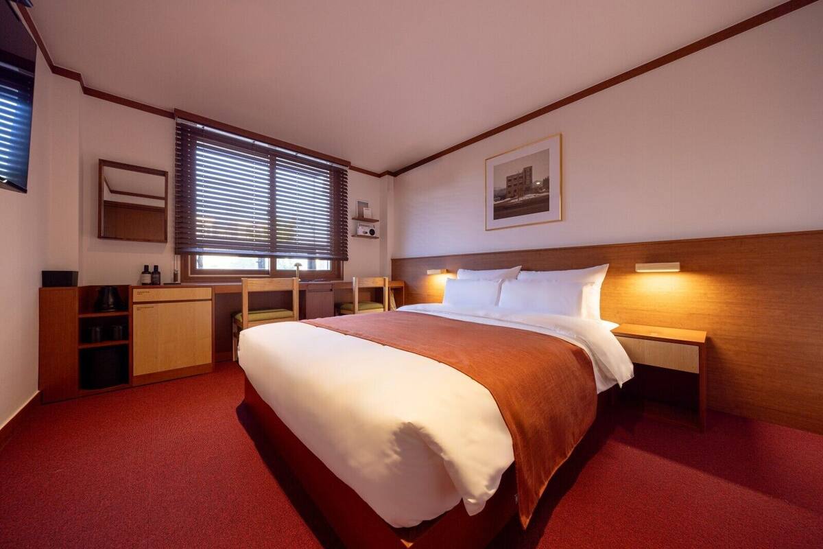 A hotel room with a large bed, a desk with a chair, a window with blinds, a bedside table with a lamp, and a framed picture on the wall.