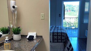Une salle de bain équipée d’un lavabo, d’un miroir, d’un sèche-cheveux et d’articles de toilette. Une chambre à coucher avec un lit et un balcon est visible à travers une porte ouverte.