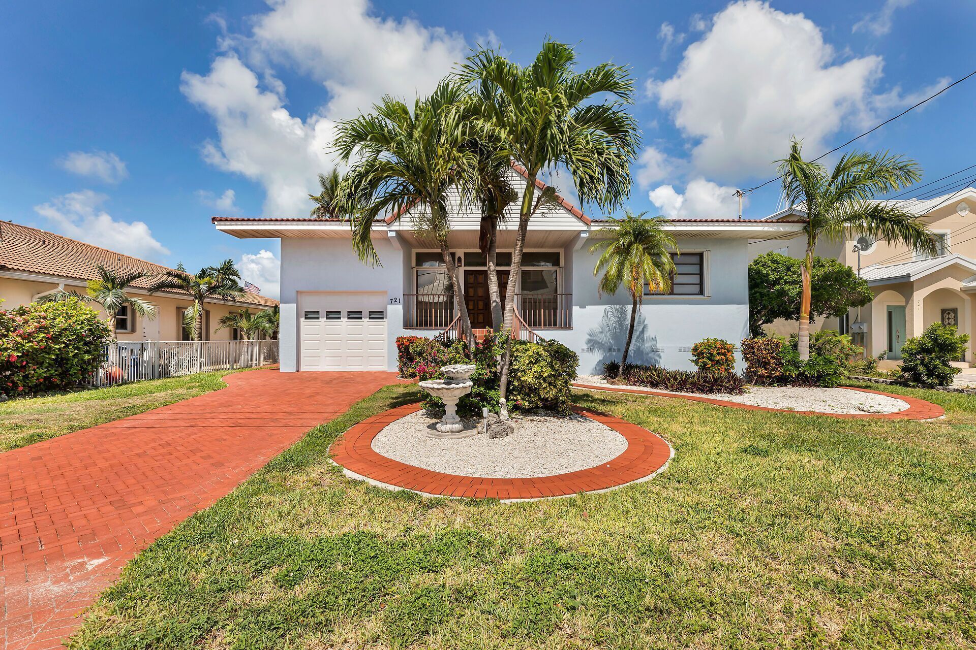 A single-story house with a white exterior, a red brick driveway, and a well-maintained lawn with palm trees.