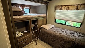 Ein kompaktes Wohnmobil-Schlafzimmer mit Etagenbetten, einer kleinen Kommode und einem Fenster mit Vorhängen.