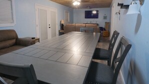 Een eetgedeelte met een lange tafel en stoelen, een bank, een televisie en een plafondventilator.
