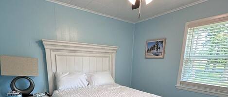 Un dormitorio con cama blanca, mesita de noche, ventilador de techo y ventana con persianas.