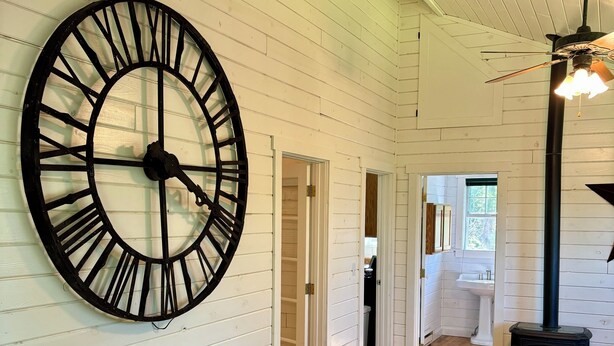 A spacious room with a large wall clock, wooden flooring, a patterned rug, and a fireplace.