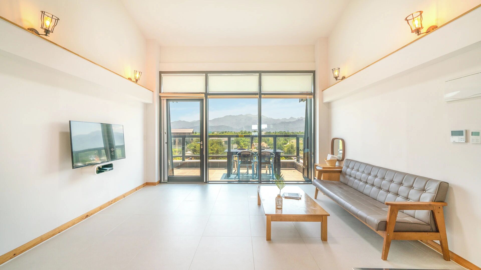 A modern living room with a flat-screen TV, a sofa, a coffee table, and a balcony with a view of the mountains.