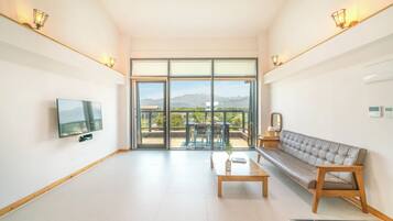 A modern living room with a flat-screen TV, a sofa, a coffee table, and a balcony with a view of the mountains.