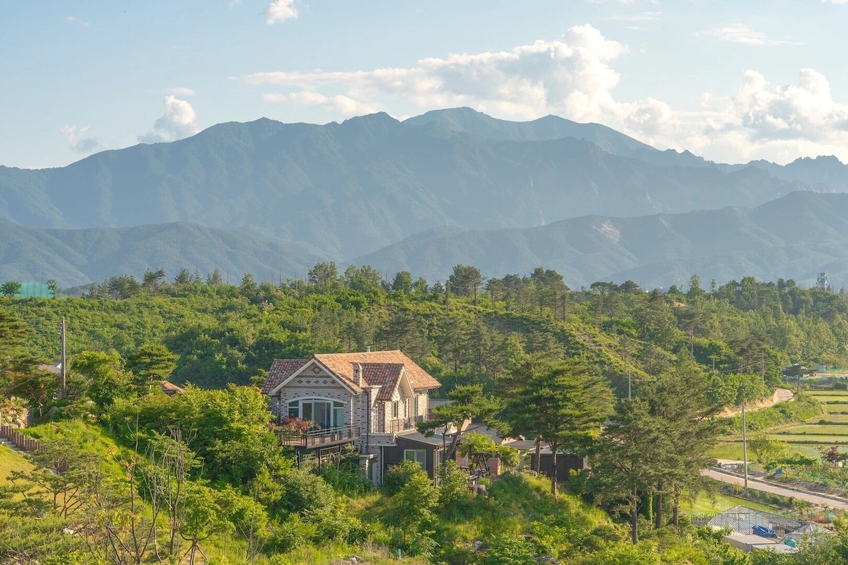 Une maison avec un toit en pente et une façade en briques, entourée d’une végétation luxuriante et avec des montagnes en arrière-plan.