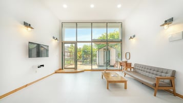 Un salón amplio con un gran ventanal corredera que da a un espacio exterior, un televisor de pantalla plana montado en la pared, un sofá, una mesa de centro y dos mesitas auxiliares.