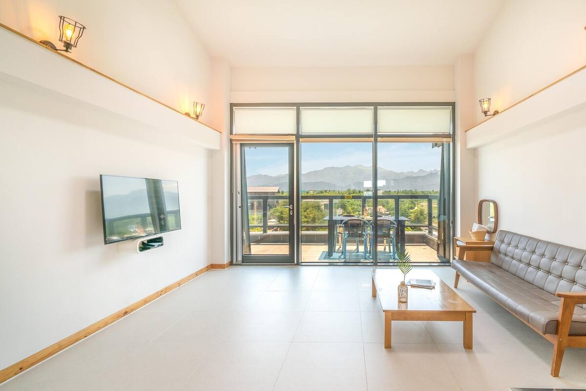 Ein geräumiges Wohnzimmer mit großen Schiebeglastüren, die auf eine Terrasse im Freien führen, einem wandmontierten Flachbildfernseher, einem Holztisch und einem grauen Sofa.