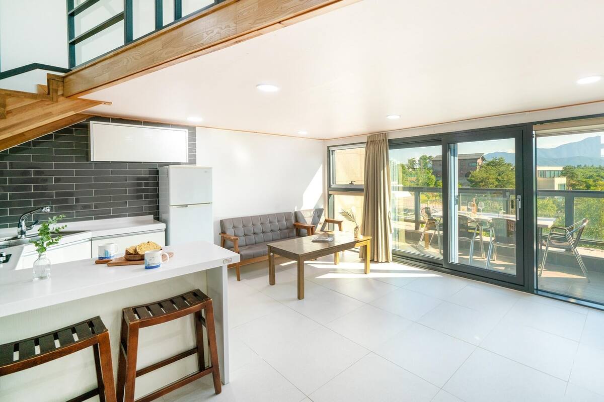 Eine moderne Küche mit Einbauofen, ein Essbereich mit einer Couch und eine große Schiebeglastür, die auf einen Balkon mit Blick auf Grünflächen und Gebäude führt.