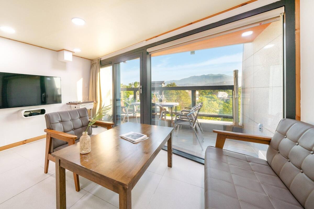 Ein modernes Wohnzimmer mit einem Holzespeisetisch, grau gepolsterten Sesseln, einem flachbildfernseher an der Wand und einem Balkon mit Blick auf Grünflächen und Berge.