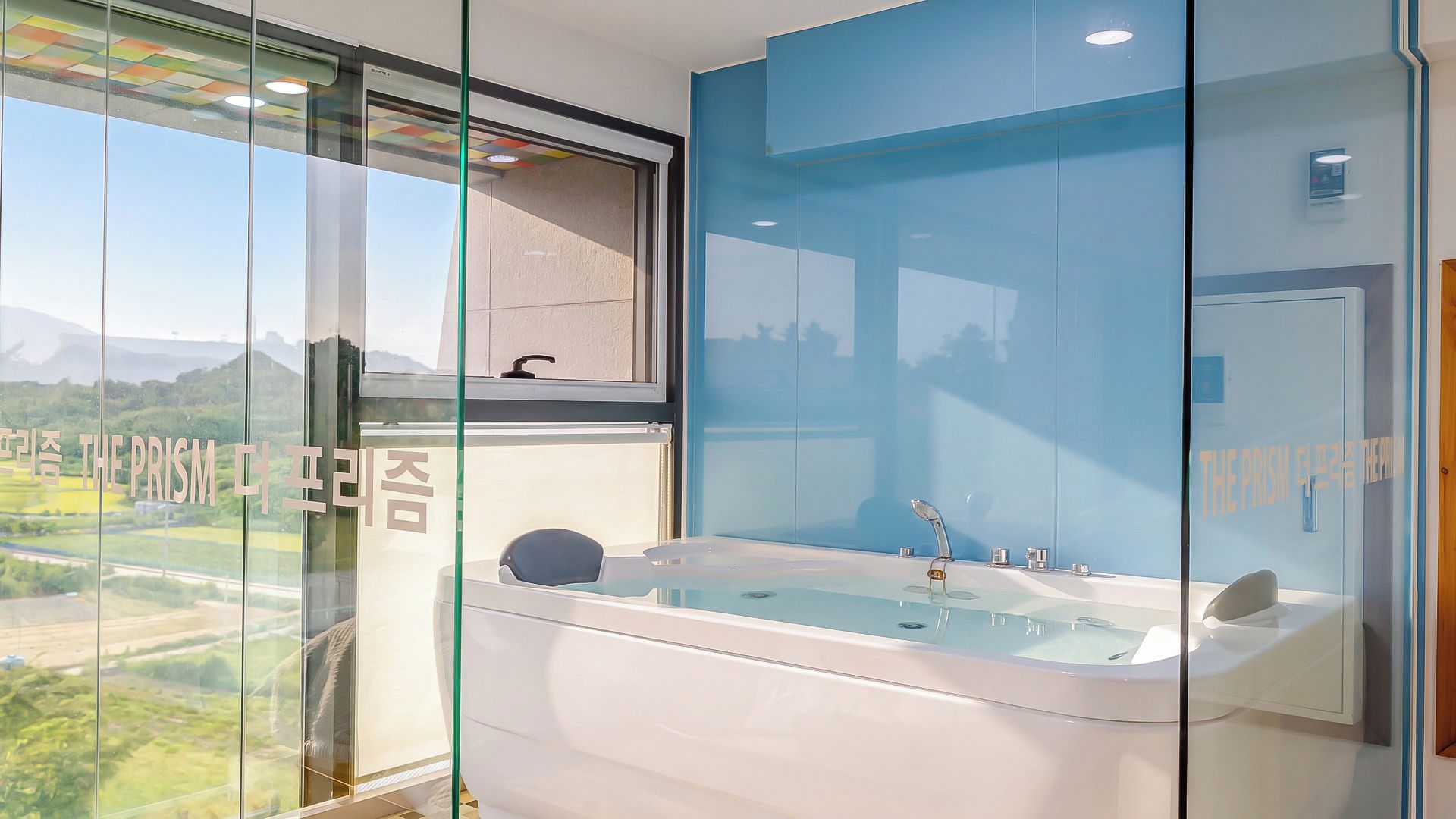 A modern bathroom with a bathtub, a window with a view of the countryside, and a glass partition with text.