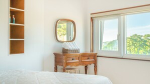 Une chambre à coucher comprenant un lit, une commode avec un miroir, une fenêtre donnant sur des arbres et une étagère avec des figurines.