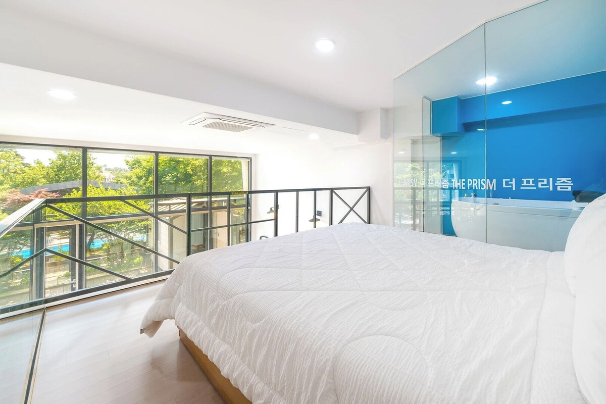 Une chambre moderne avec un grand lit, une cloison en verre et une vue sur la verdure environnante.
