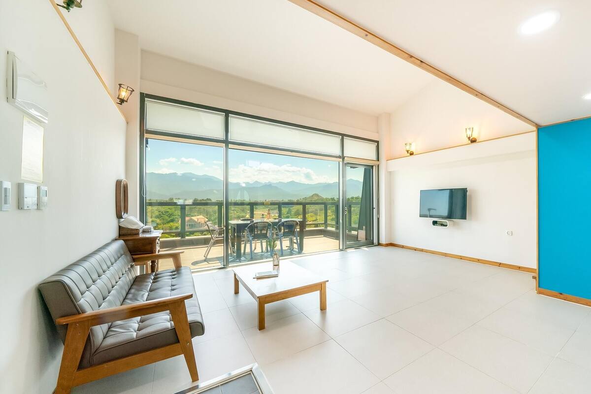 Ein geräumiges Zimmer mit einer großen Schiebeglasdoor, die auf einen Balkon mit Bergblick führt. Es gibt ein Sofa, einen Couchtisch und einen an der Wand befestigten Fernseher.