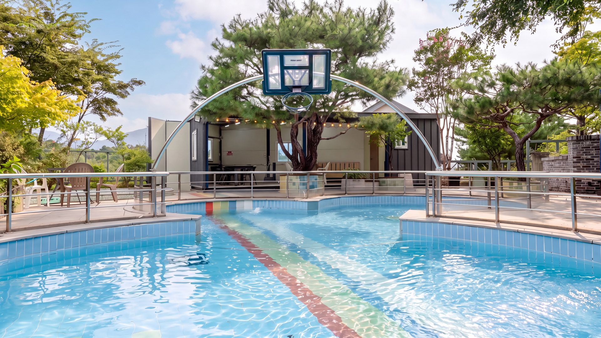 A swimming pool with a basketball hoop, surrounded by trees and a building with outdoor seating.