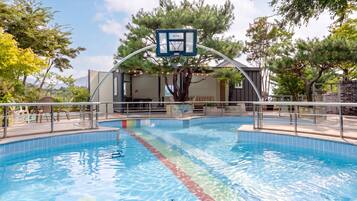 A swimming pool with a basketball hoop, surrounded by trees and a building with outdoor seating.