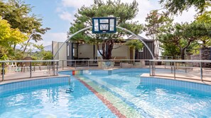 A swimming pool with a basketball hoop, surrounded by trees and a building with outdoor seating.