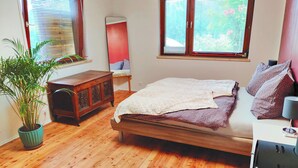 A bedroom with a wooden floor, a bed with a white blanket, a wooden dresser, a potted plant, and two windows with blinds.
