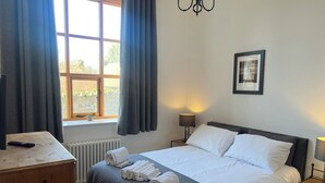 A neatly made bed with white linens and rolled towels, a bedside table with a lamp, a wooden dresser, and a window with curtains. - Barnstaple - Apartment- Sleeps 2 - Parking (Barnstaple)