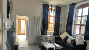 A living room with a sofa, a coffee table, a small table with chairs, a radiator, and a window with curtains. - Barnstaple - Apartment- Sleeps 2 - Parking (Barnstaple)