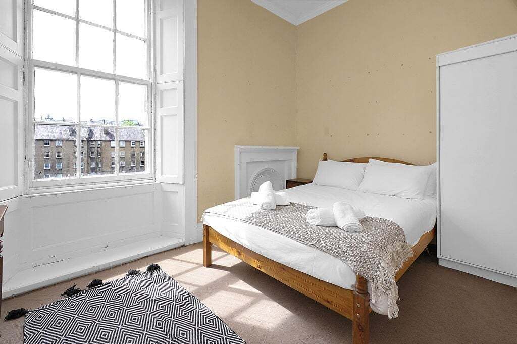 A bedroom with a large window, a bed with white linens, a radiator, and a patterned ottoman.