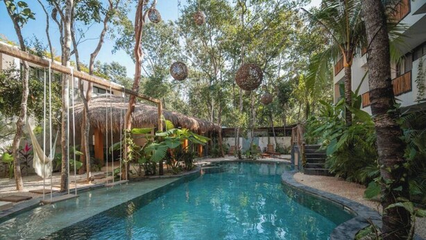 A swimming pool surrounded by tropical vegetation, a thatched-roof structure, and a multi-story building with balconies. - Tulum Getaway+Pool+Office Area+Gym (Tulum)