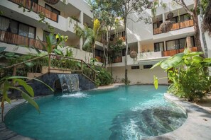 Ein Pool mit Wasserfall, umgeben von üppiger Bepflanzung und einem mehrstöckigen Gebäude mit Balkonen.