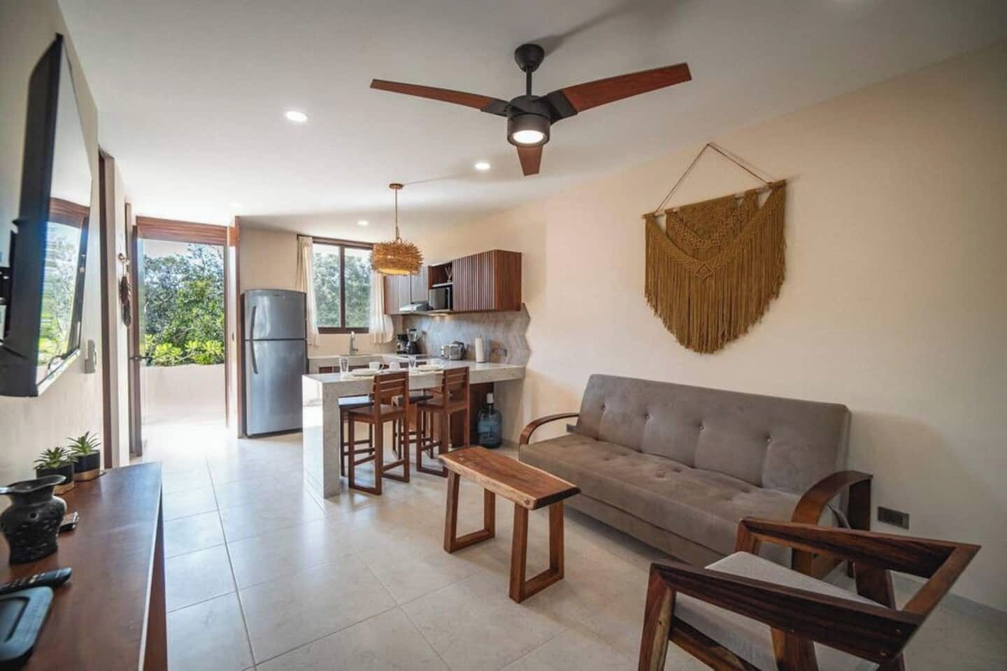A modern living room with a sofa, a dining area with chairs, a kitchen with a refrigerator, and a ceiling fan.