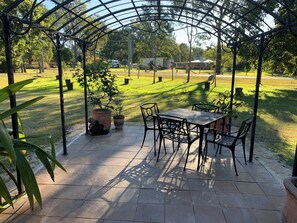 Un porche cubierto con una mesa y sillas, rodeado de plantas en macetas y un césped.