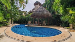 Una piscina con un gazebo a tetto di paglia, circondata da palme e poltroncine da relax.