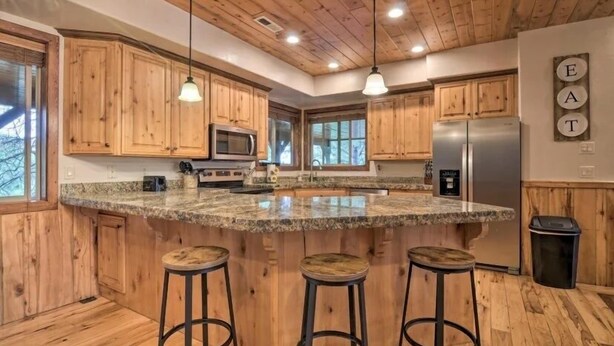A kitchen with wooden cabinets, granite countertops, a central island with stools, stainless steel appliances, and pendant lighting. - Bridgeway House (Duck Creek Village)